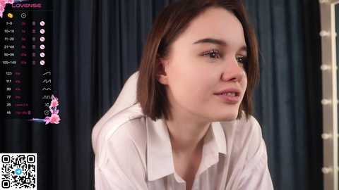 Media: Video of a young woman with shoulder-length brown hair, wearing a white blouse, seated indoors with a dark curtain background. She gazes thoughtfully to the side.