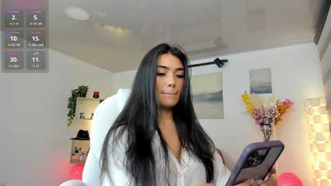 Media: Video of a young woman with long black hair, fair skin, and medium-sized breasts, wearing a white robe, playing on a tablet in a modern, minimalist bedroom with white walls, a potted plant, and a window.