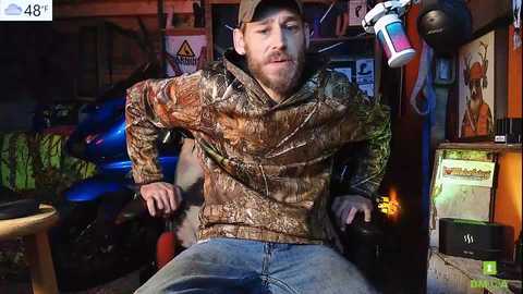 Media: Video of a bearded man in a camo jacket and jeans, sitting in a cluttered workshop. Background includes tools, a motorcycle, and various objects, creating a rustic, industrial atmosphere.