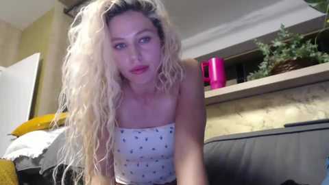 Media: A video of a blonde woman with long, curly hair, wearing a white spaghetti-strap tank top with small black designs, leaning forward on a dark couch, in a cozy living room with a pink kettle, plants, and a yellow blanket.