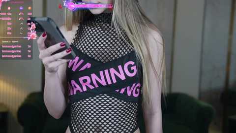 Media: Video of a slender, light-skinned woman with long blonde hair, wearing a black fishnet top with \"WOMAN\" in pink letters. She holds a smartphone with a pink light filter. Background shows a dimly lit room with a green couch.