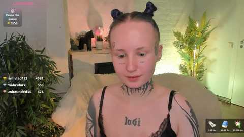 Media: A young woman with pale skin, black hair in two buns, and a black lace top sits indoors. She has tattoos on her neck and arms, and the room has a green plant, a lamp, and a white wall.