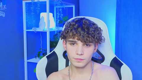 Media: Video of a young, shirtless white boy with curly brown hair, seated in a white gaming chair, in a brightly lit room with blue lighting.