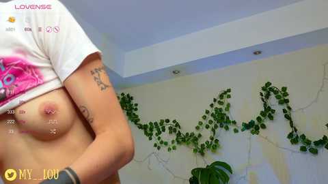 Media: A topless woman with medium-sized breasts, visible nipple piercings, and tattoos on her arms stands in a bedroom with green vines on the wall and a plant in the corner.