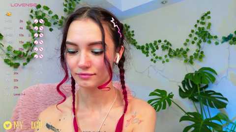 Media: Video of a young woman with fair skin, dark hair in braids, and a red ribbon, wearing a red top, standing in a greenery-filled room with a calendar and plants in the background.