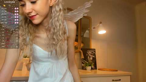 Media: A video of a young woman with long, wavy blonde hair, wearing a white, strapless dress, and angel wings, standing in a cozy, dimly-lit room with a wooden vanity and a wall calendar.