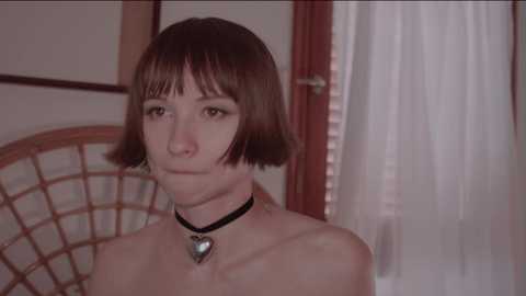 Media: Video of a young woman with short brown hair, wearing a black choker with a heart pendant, topless, in a dimly lit room with wooden furniture and a white curtain.