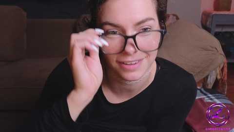 Media: Video of a smiling, fair-skinned woman with curly dark hair, wearing black-rimmed glasses, adjusting them, in a cozy living room with a brown sofa and plaid blanket.