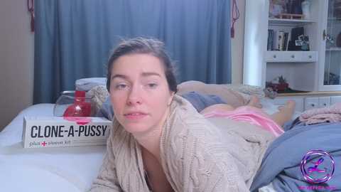 Media: Video of a woman in a beige knit cardigan lying on a bed, reading \"Cloned-A-Pussy\" book, blue curtains, messy room.