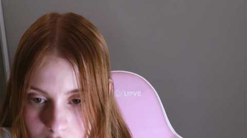 Media: Video of a young woman with long, straight, reddish-brown hair, sitting in a pink chair with a white backrest. She has light skin and is gazing downward. The background is a plain, light gray wall.