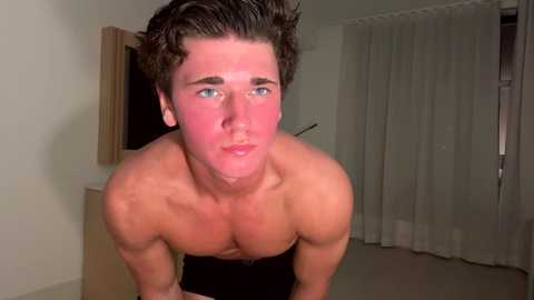 Media: Video of a shirtless, muscular young man with tanned skin, dark curly hair, and a red sunburn on his face, standing indoors against white walls and sheer curtains.