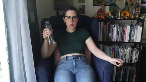 Media: A woman with glasses and a green shirt sits in a dark blue chair, holding a water bottle. Bookshelves and sunflowers are in the background.