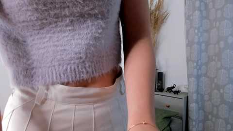 Media: Video of a person in a gray, ribbed, cropped sweater and white pleated skirt, holding a phone. Background includes a white wall, patterned curtain, and a small, cluttered white dresser.