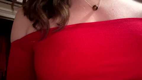 Media: A close-up video of a woman in a bright red off-the-shoulder dress, showing her bare shoulder and a necklace with a brown pendant. Her wavy brown hair falls over her left shoulder.