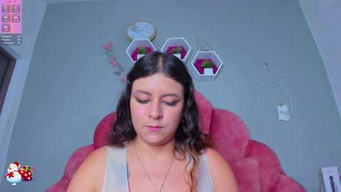 Media: Video of a plus-size woman with long dark hair, wearing a white tank top, sitting on a pink, plush chair. Background features a green wall with a dreamcatcher, hexagonal plant holders, and a pink heart.