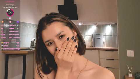 Media: Video of a young woman with shoulder-length brown hair, covering her mouth with her hand, in a modern kitchen with tiled backsplash, visible chat box in the corner.