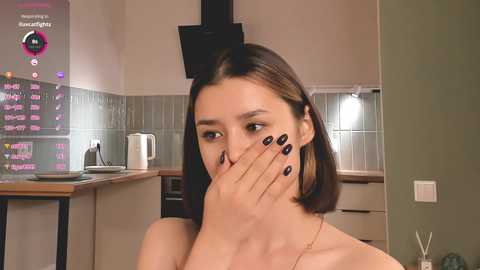 Media: A young woman with shoulder-length brown hair, wearing black nail polish, covers her mouth in a modern kitchen with tiled backsplash, digital screen displaying stock market data.