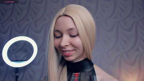 Media: Video of a light-skinned woman with straight, long blonde hair, wearing a black leather-like top with a plaid collar, smiling softly. Background features a gray, floral-patterned wall and a ring light to her left.