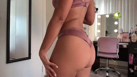 Media: Video of a woman in mauve lingerie, highlighting her toned buttocks, standing in a modern vanity room with a mirror and a pink chair.