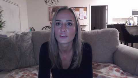 Media: Video of a blonde woman with light skin, wearing a black top, sitting on a light gray couch in a cozy living room with neutral decor and a kitchen in the background.