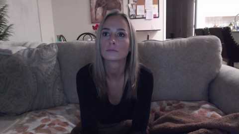 Media: Video of a young woman with long blonde hair and light skin, wearing a black top, sitting on a gray couch with patterned pillows and a brown blanket. Background includes a corkboard with pins and a window.