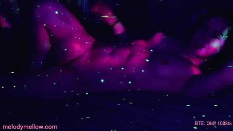 Media: Video of a nude woman with a toned physique, lying on a dark surface. The image is dimly lit with purple and pink hues, creating a sensual, ethereal ambiance.