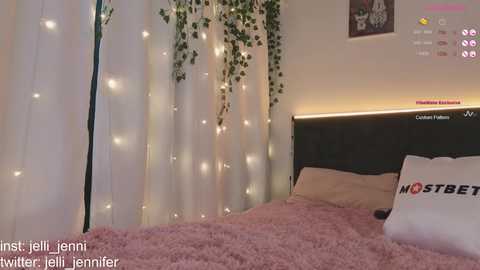 Media: Video of a cozy bedroom with a dark headboard, white curtains adorned with fairy lights, a plush pink bedspread, and a white pillow with \"HOSTES\" text.