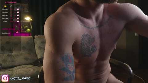 Media: Video of a shirtless, muscular man with tattoos on his chest and arm, sitting on a couch in a dimly lit living room.