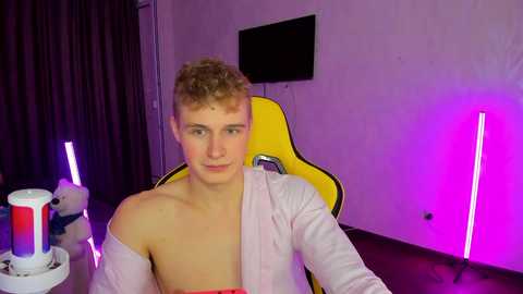 Media: Video of a young, fair-skinned, curly-haired man in a pink shirt, sitting in a yellow gaming chair. Background includes a TV, purple and blue neon lights, and a Pepsi can.