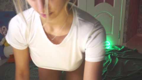 Media: A video of a blonde woman in a white t-shirt, leaning forward with her head slightly tilted. The background features a door, green lights, and a plush toy.