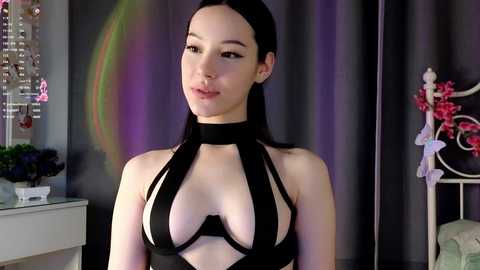 Media: Video of a young, pale-skinned woman with long black hair, wearing a revealing black halter top, standing in a dimly lit room with a rainbow backdrop, and floral decor.