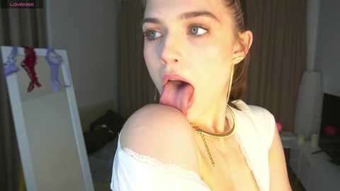 Media: Video of a fair-skinned, blue-eyed woman with long brown hair, wearing a white off-shoulder top and gold necklace, sticking her tongue out in a bedroom with a full-length mirror and clothing hanging.