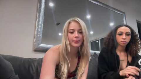 Media: Video of two young women on a gray couch. One has long blonde hair, a red dress, and a small chest. The other has curly black hair, a black jacket, and a medium chest.