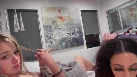 Media: Video of a young woman with blonde hair and another woman with curly brown hair, both wearing gray sweaters, in a modern bedroom with abstract art and a mirror.