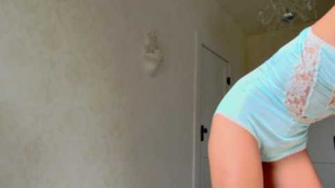 Media: Video of a woman bending over, wearing light blue lace-trimmed panties, in a sparsely furnished room with a white door and textured beige wall.