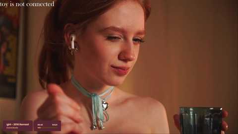 Media: Video of a fair-skinned, red-haired woman with a heart necklace, wearing headphones, holding a glass, smiling slightly, in a warm, dimly lit room.