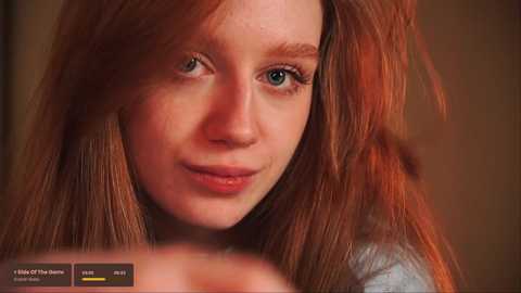 Media: Video of a fair-skinned, freckled young woman with long, tousled red hair and blue eyes, wearing a gray top, smiling gently. Background is blurred.