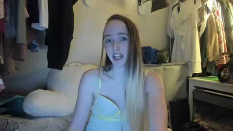 Media: Video of a young, light-skinned woman with long, straight blonde hair wearing a yellow bra, sitting on a bed in a messy bedroom with clothes hanging on a rack and a dresser.