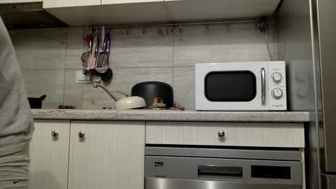 Media: Video of a minimalist kitchen with a beige countertop, white cabinets, a black kettle, a microwave, and utensils hanging from a rack.