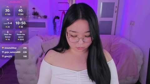 Media: Video of a young Asian woman with long black hair, wearing glasses and a white off-shoulder top, sitting on a plush couch, surrounded by purple lighting, with a virtual reality overlay displaying chat messages.