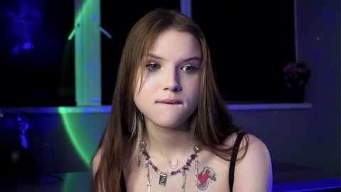 Media: Video of a young Caucasian woman with long brown hair, wearing a black top, silver necklace, and tattoos, in a dimly lit room with neon lights.