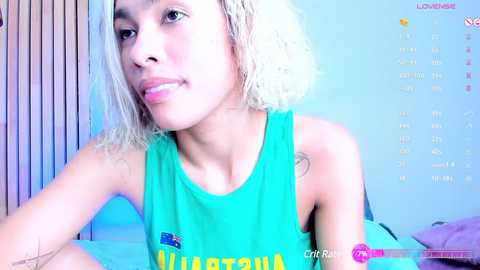 Media: Video of a light-skinned woman with short, platinum blonde hair, wearing a green tank top with \"Australia\" in yellow. She has a soft smile and a youthful appearance. Background includes a calendar, a bed, and a vertical window blind.