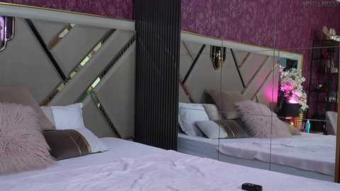 Media: Video of a luxurious bedroom with a plush white bed, a large mirror headboard, and a plush pink pillow. The walls are adorned with intricate floral wallpaper, and a black vertical column separates the bed from the mirror.