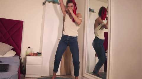 Media: A video of a woman in a white tank top and blue jeans, striking a pose in front of a mirror in a modest bedroom.