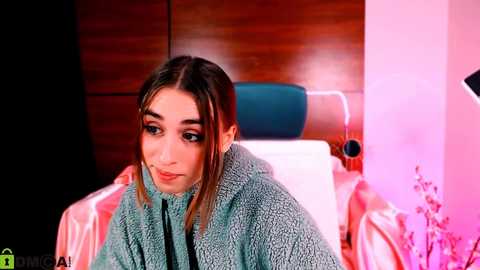 Media: Video of a young woman with light skin, brown hair, and green eyes, wearing a blue fleece jacket, seated in a modern, brightly lit room with a wooden headboard and pink lighting.