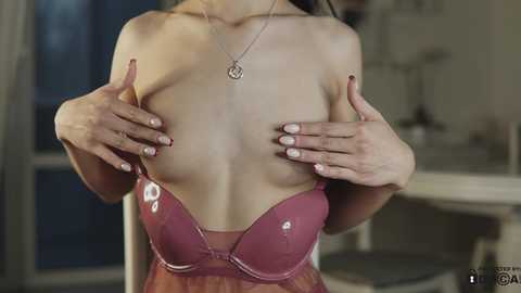 Media: Video of a woman with fair skin and long dark hair, wearing a burgundy lace bra, holding her breasts, and a silver necklace.