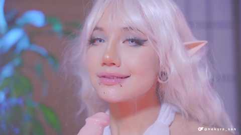 Media: Video of a pale-skinned, slender woman with long, platinum blonde hair, elf-like pointed ears, and light makeup. She wears a sleeveless white top, smiling with her lips slightly parted. Background features a blurred green plant and a window.