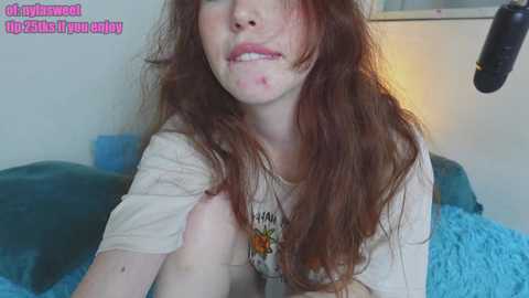 Media: Video of a young woman with long, reddish-brown hair and pale skin, wearing a white t-shirt, with a red mark on her cheek. She's lying on a bed with a blue blanket.