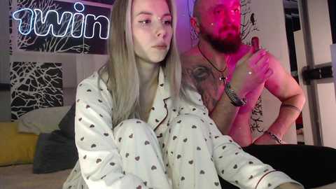 Media: Video of a blonde woman in pajamas and a bearded man in a tank top, both in a dimly-lit room with neon lights and abstract art.