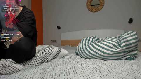 Media: Video of a woman in black pants and a dark top, lying on a bed with white and green striped sheets, watching a game on a TV in a modern bedroom.